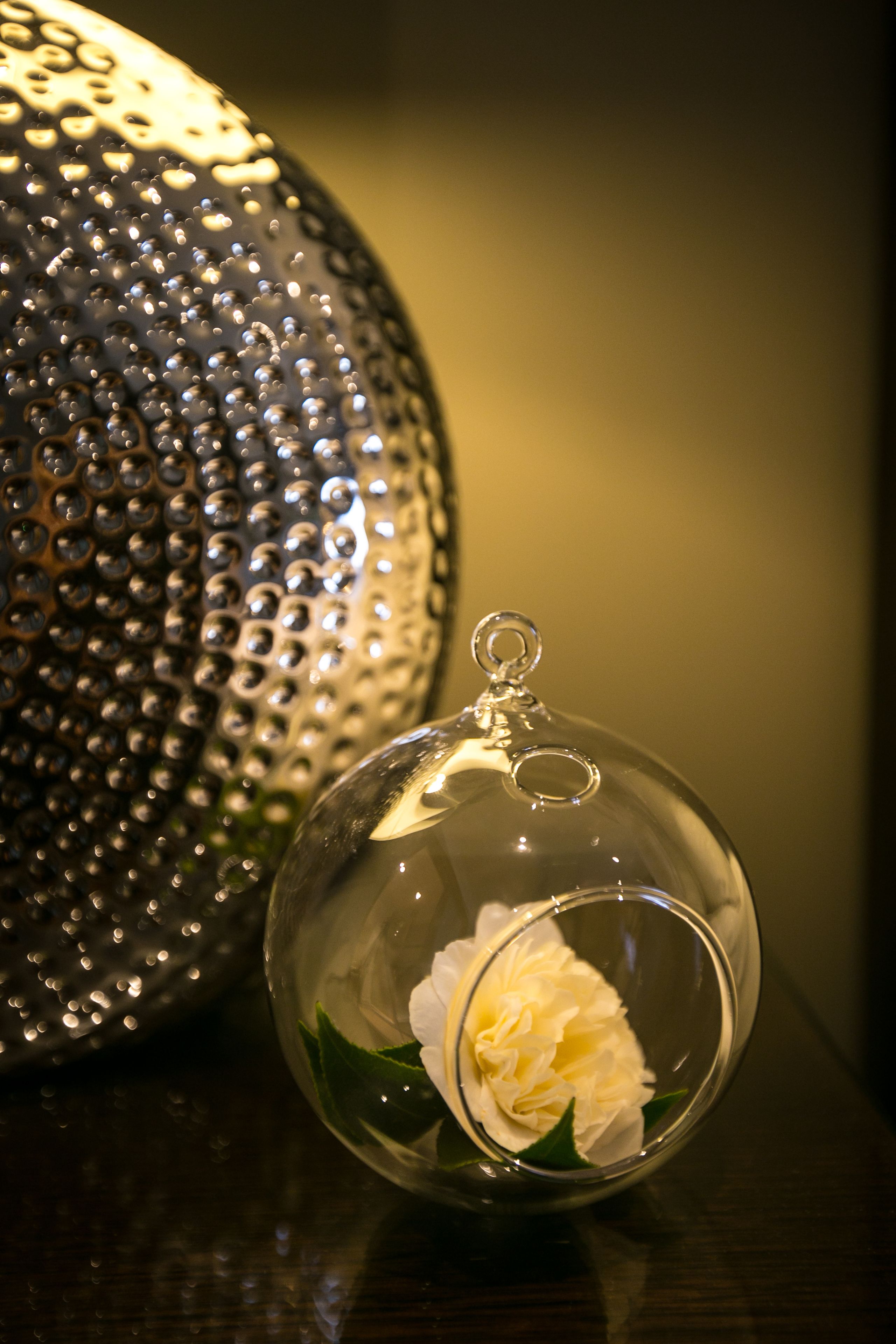 A clear glass ornament with a white flower and a metallic textured sphere in the background.