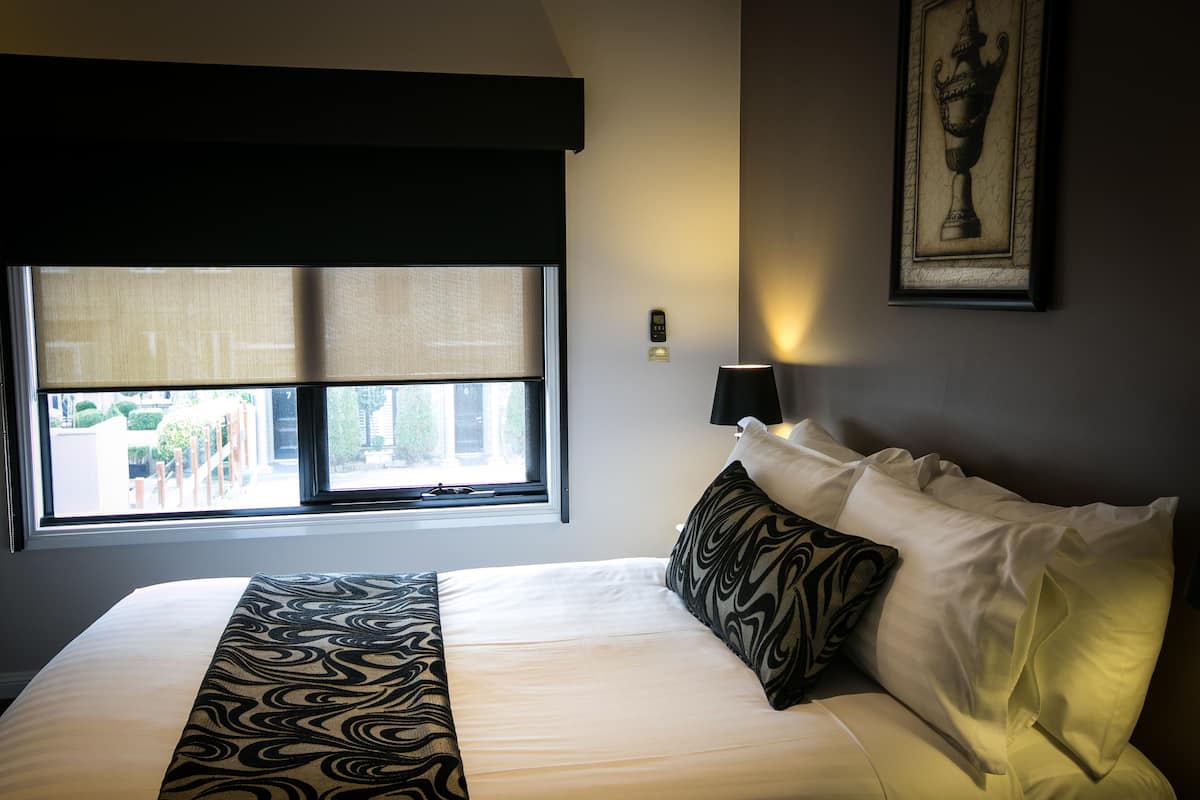 Una cama bien tendida con una colcha estampada, un ventanal con vistas, una lámpara de pared y un cuadro enmarcado.