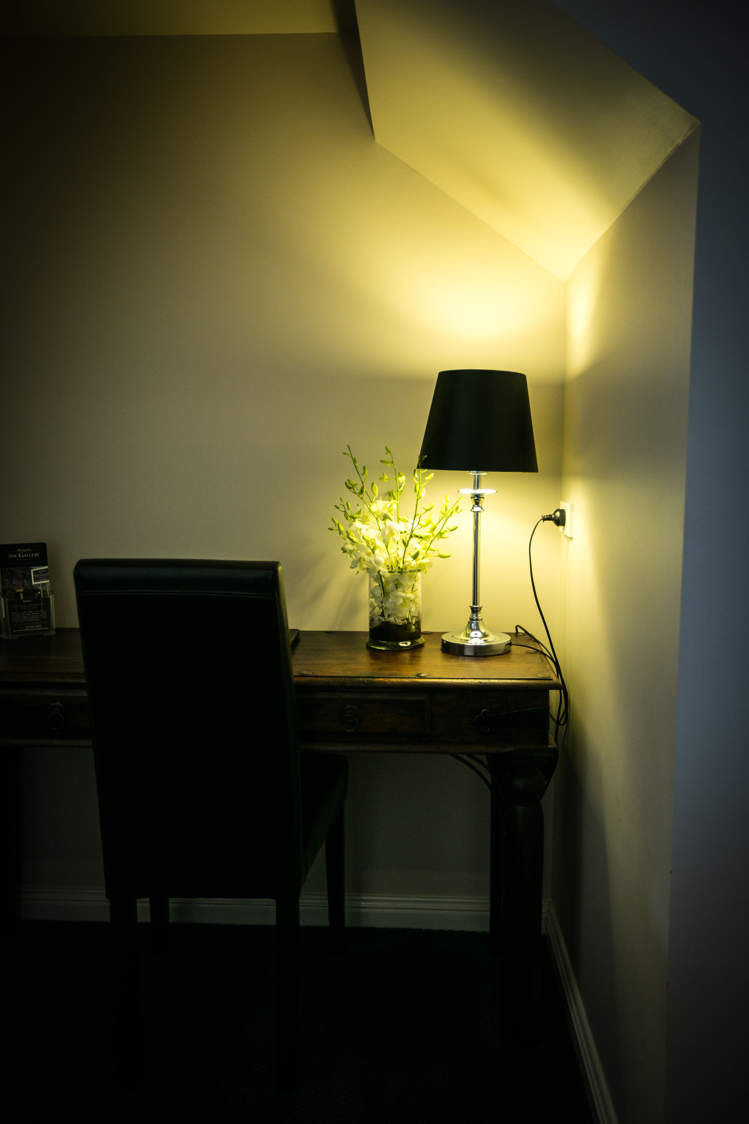 Un escritorio con lámpara y un jarrón de flores, una silla y un enchufe de pared.