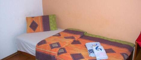 A small, single-bed room with a plain white wall and a beige floor. There is a bed with a patterned orange and brown bedspread and a single pillow with a geometric design.