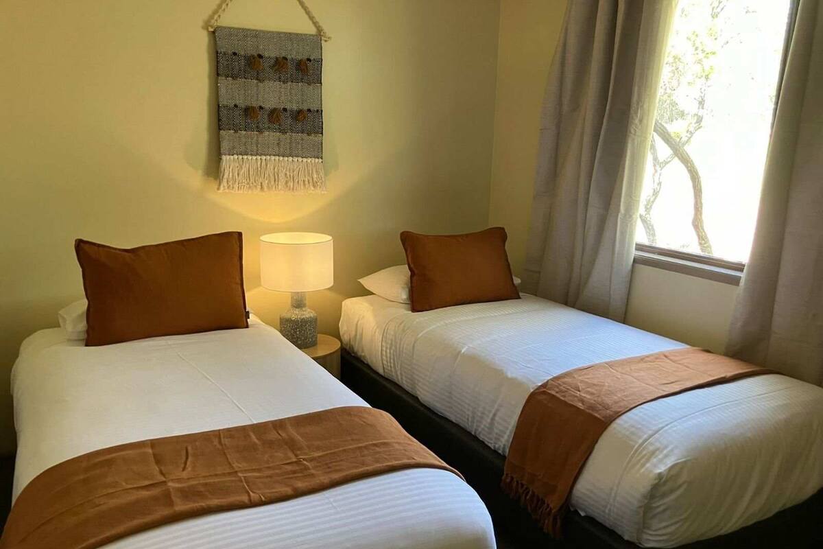 Habitación de hotel con dos camas individuales, cada una con un cojín marrón y una colcha blanca. Hay una mesita de noche con una lámpara entre las camas, y un tapiz en la pared sobre la cama de la izquierda.