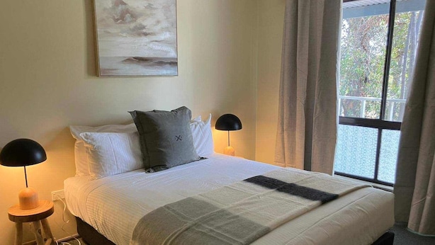 Un lit bien fait, avec du linge de lit blanc et un plaid gris, deux lampes de chevet, un tableau au-dessus du lit et une fenêtre avec des rideaux.