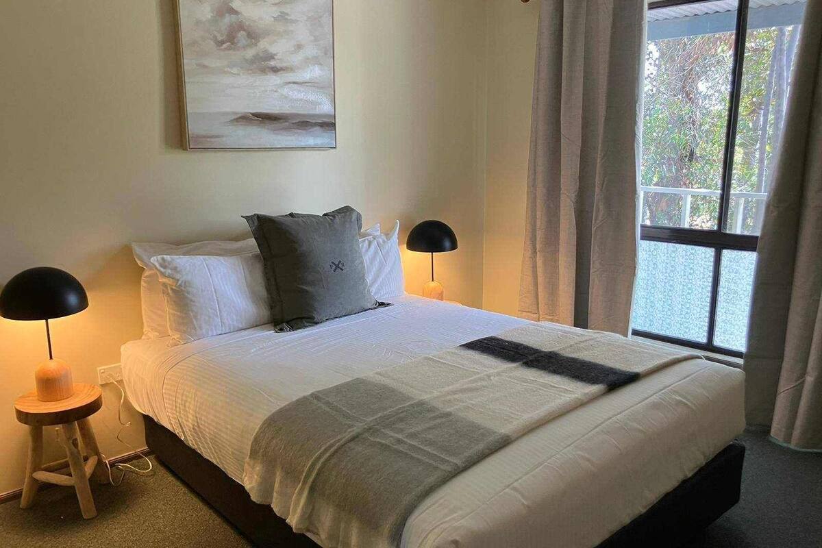 Una cama bien tendida con sábanas blancas y un plaid gris, dos lámparas de noche, un cuadro sobre la cama y una ventana con cortinas.