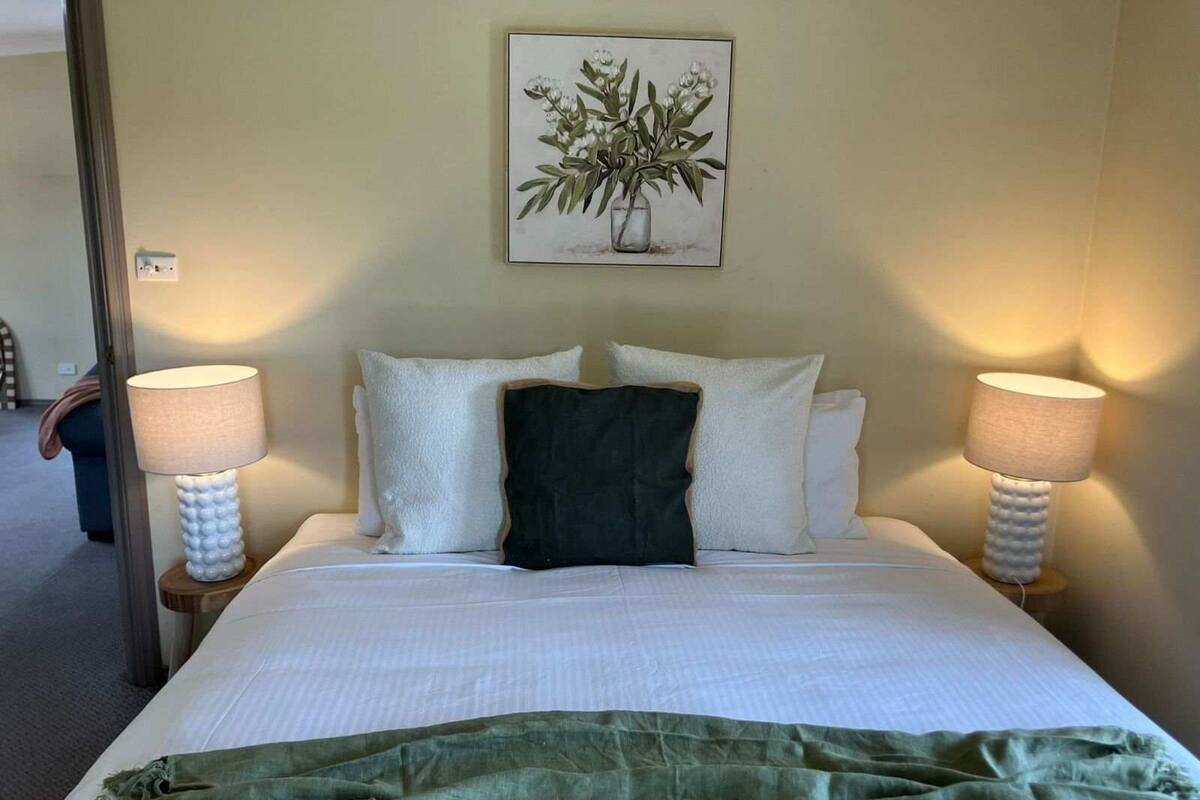 Una cama bien tendida con ropa blanca y un edredón verde, flanqueada por dos lámparas de noche y un cuadro de flores colgado en la pared.