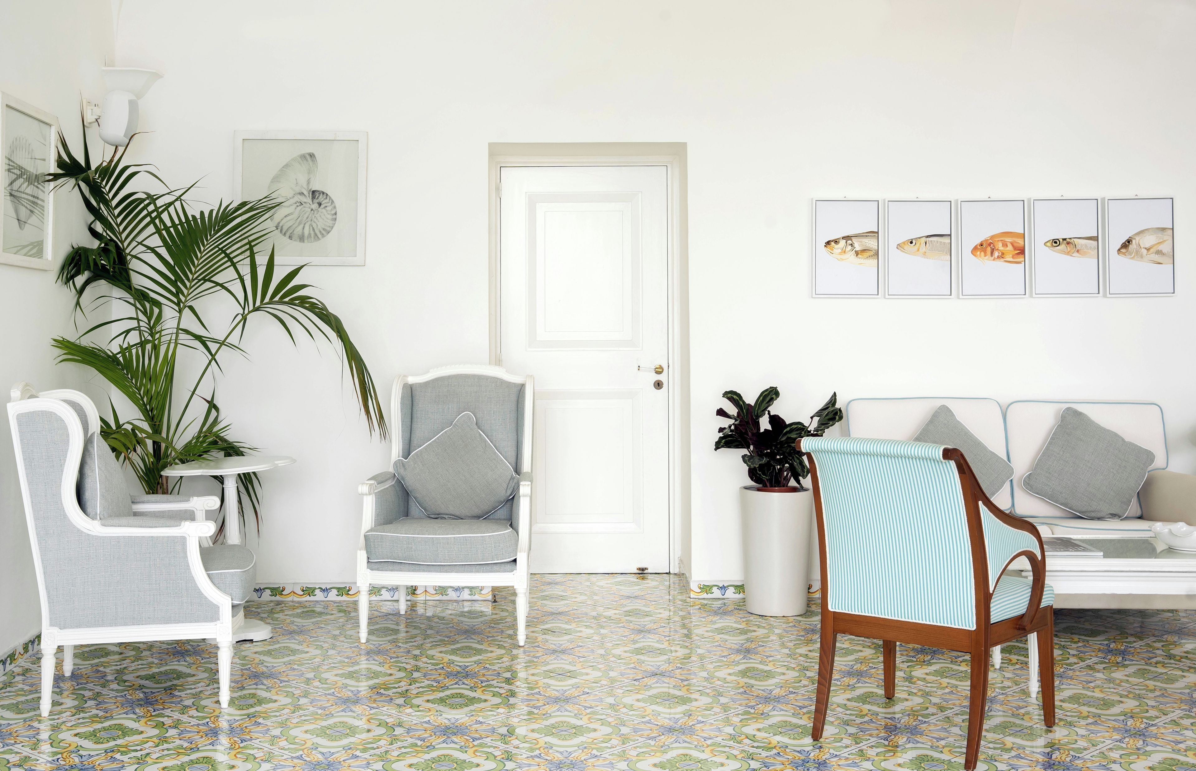 Een kamer met een witte deur, een witte bank, een gestreepte stoel en een tafel met stoelen. Er hangen fotolijsten aan de muur.