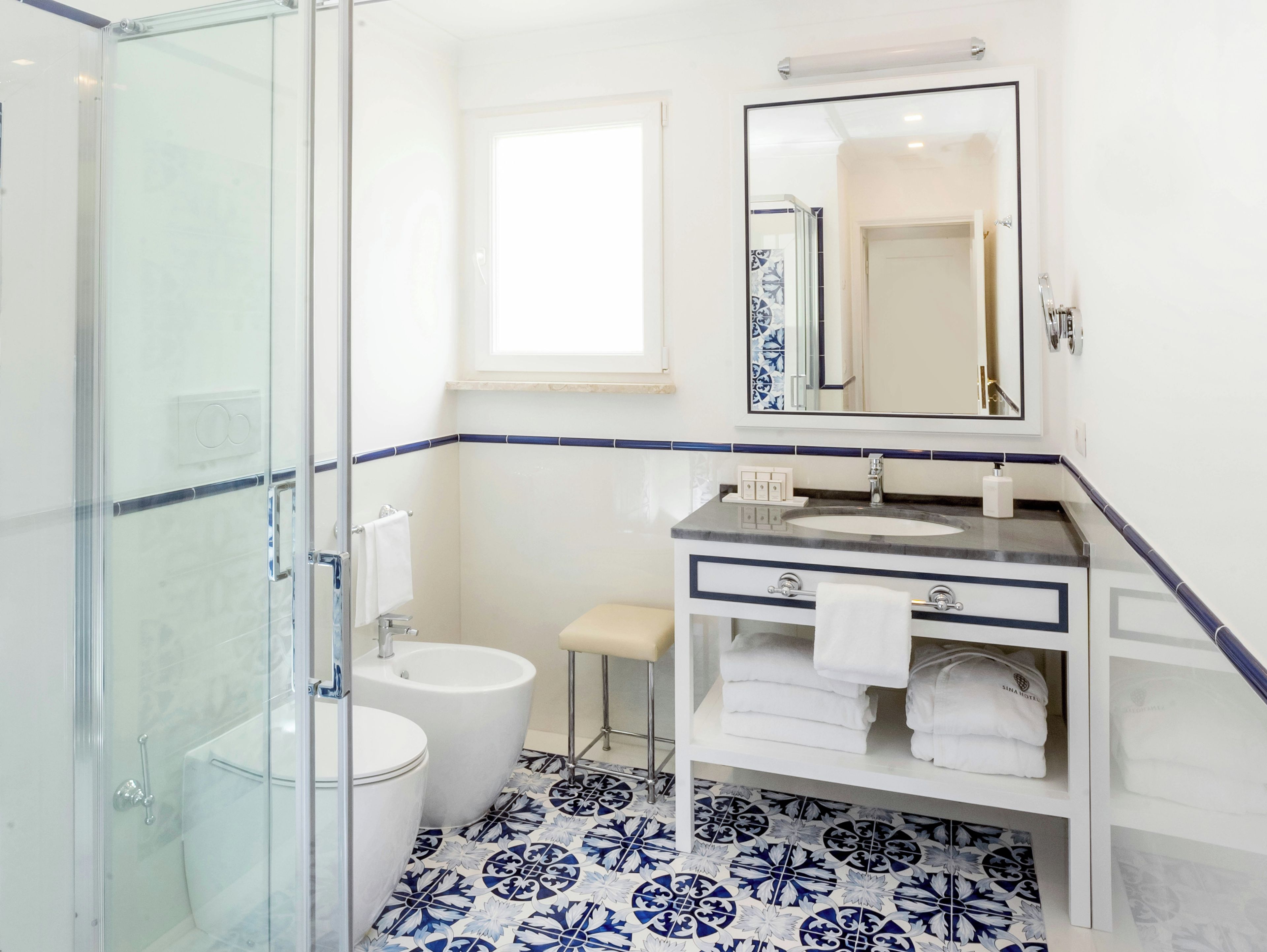 Un bagno con cabina doccia in vetro, un water bianco, un lavandino con specchio sopra e piastrelle a motivi sul pavimento.