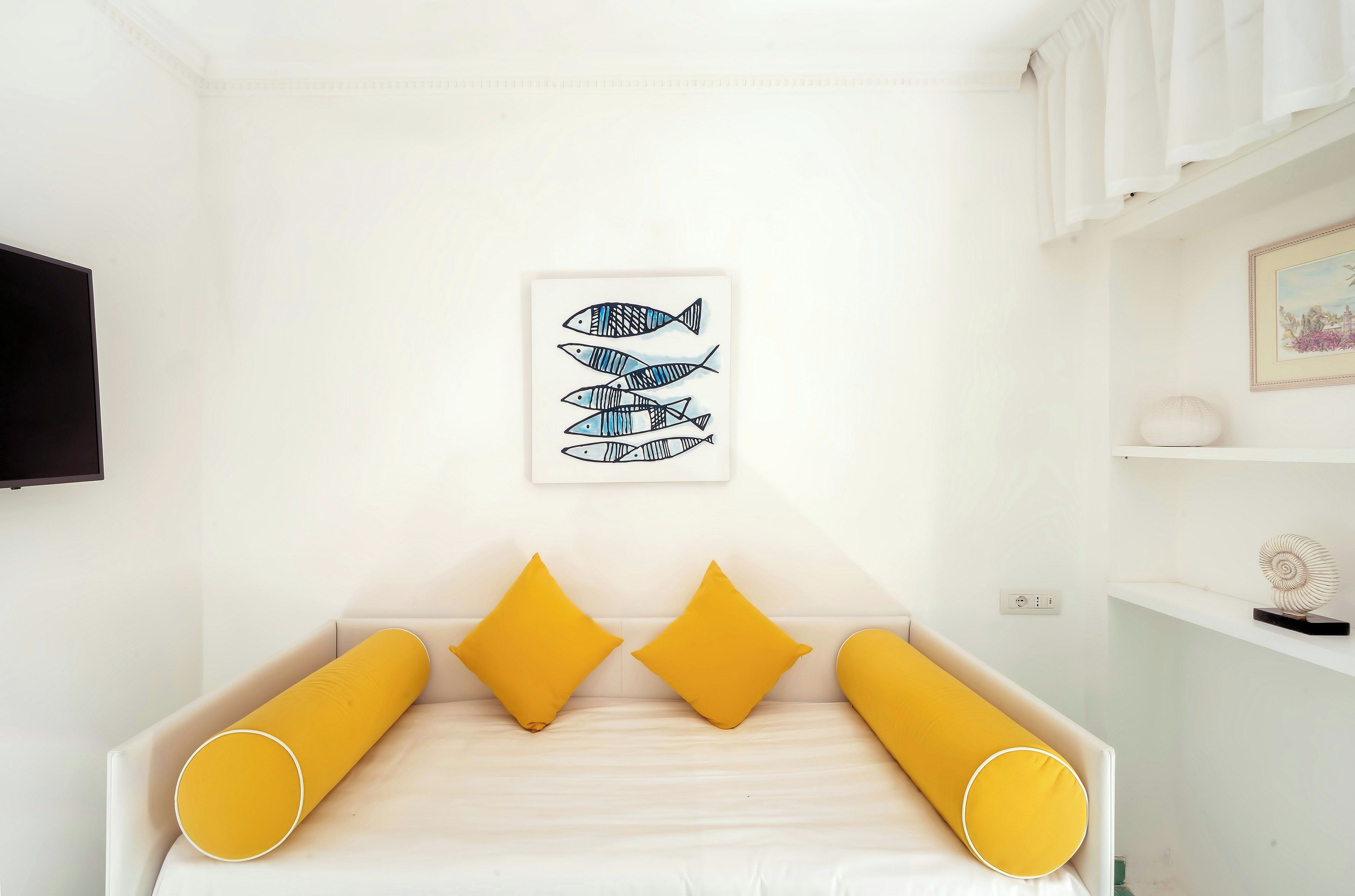 Quarto com cama, almofadas amarelas, uma obra de arte de peixes na parede e uma prateleira com objetos decorativos.