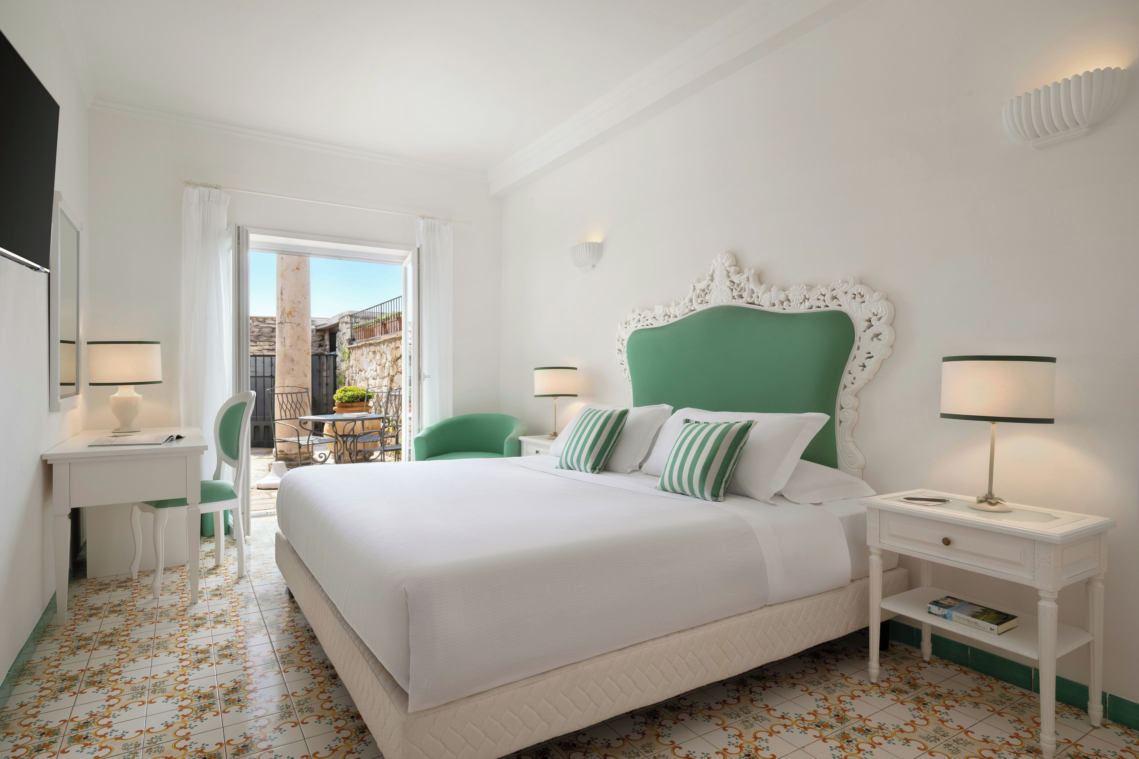 A hotel room with a bed, a desk, a chair, a nightstand, and a view of a building outside.