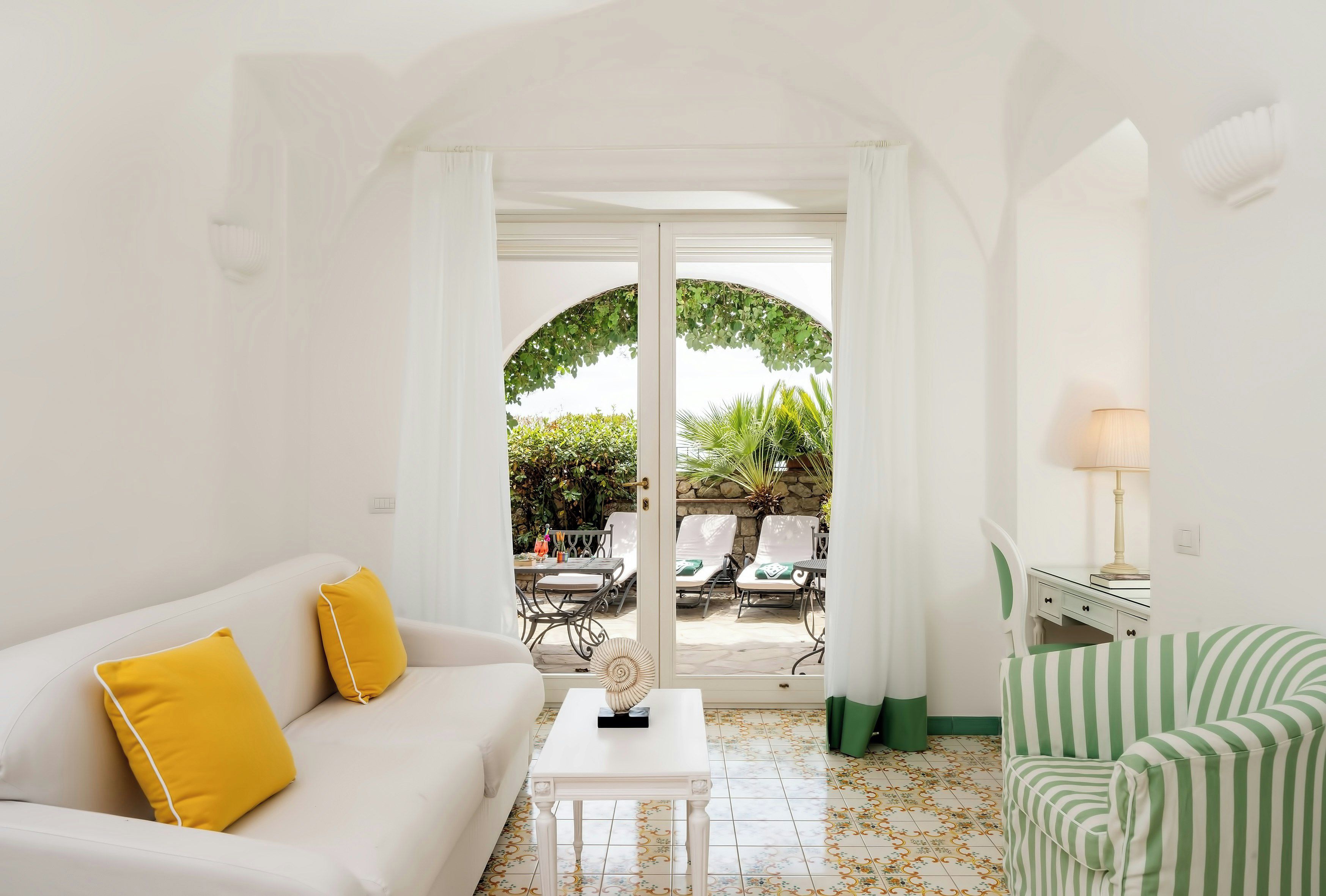 A room with a white sofa, a striped chair, a small table, and a view of an outdoor patio with chairs and a table.