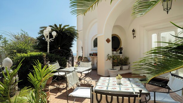 A patio with outdoor seating, potted plants, and a white building with arches. - Sina Flora Capri (Capri)