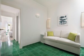 Ein Zimmer mit grün gefliestem Boden, einem weißen Sofa mit grünen Kissen, einer Wandleuchte und einem Kunstwerk zum Thema Fische.