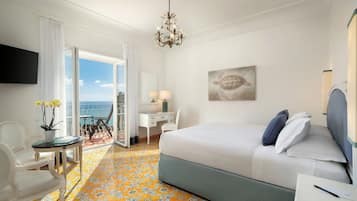 A hotel room with a bed, a desk, a chair, a TV, a chandelier, and a view of the sea.