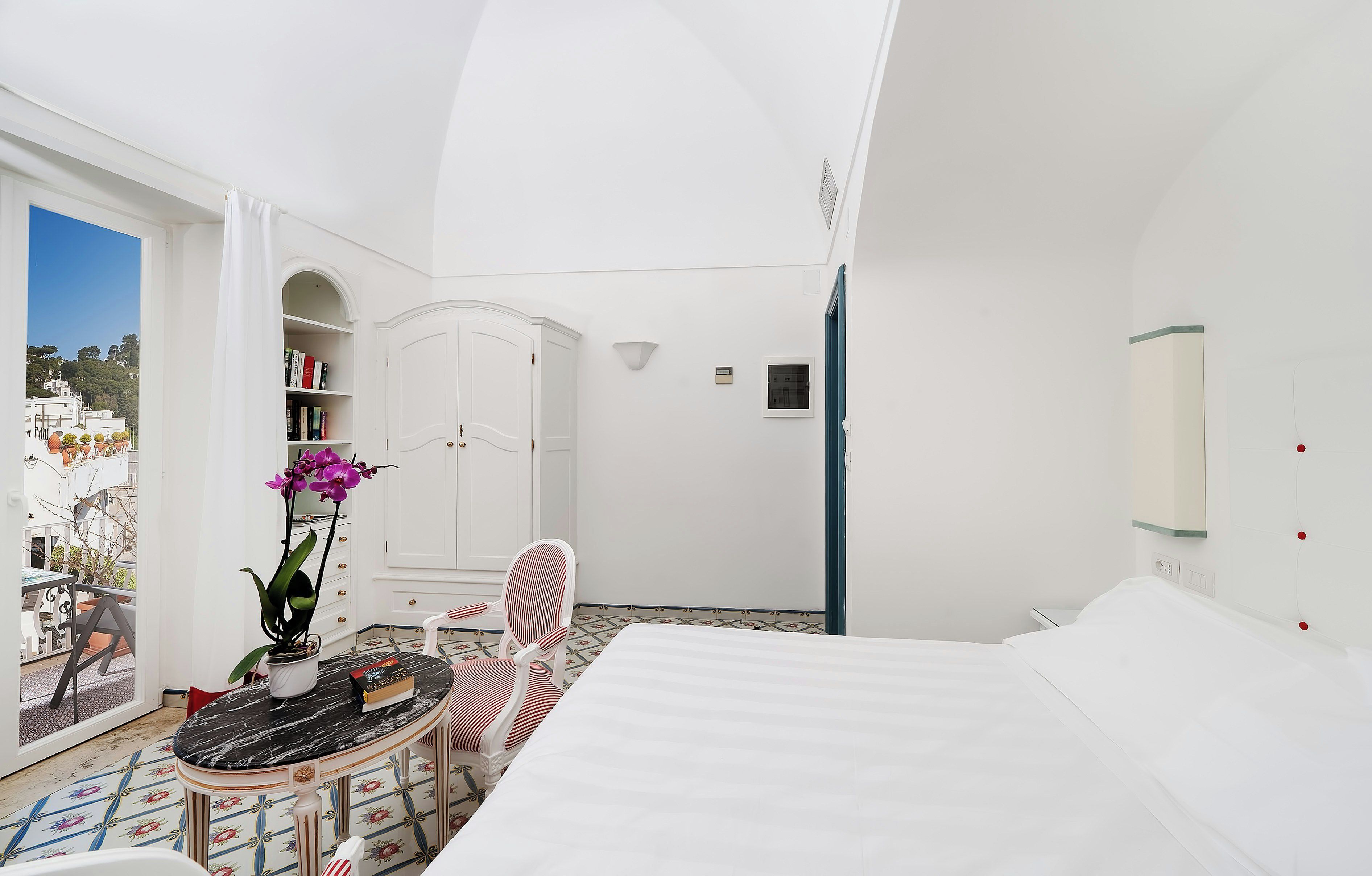 Habitación blanca con una cama, una mesita con una planta, una silla, una estantería y vistas al exterior.