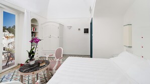 Habitación blanca con una cama, una mesita con una planta, una silla, una estantería y vistas al exterior.