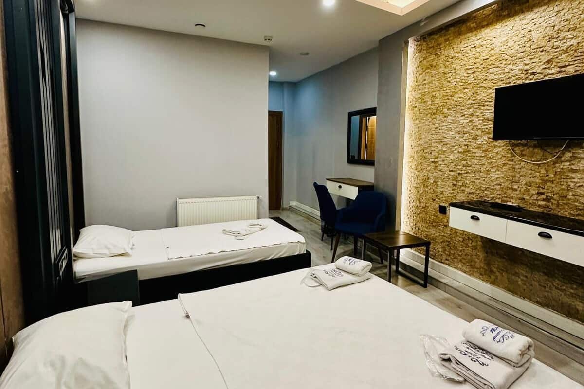 A hotel room with two beds, a desk, a chair, a TV mounted on a textured wall, and a ceiling light.