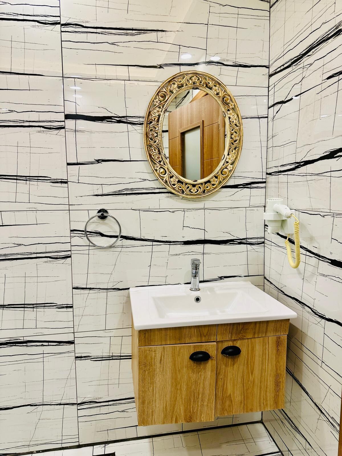 Une salle de bain avec un mur en marbre blanc, un meuble en bois doté de deux tiroirs, un lavabo avec un robinet, un miroir rond avec un cadre orné et un distributeur de serviettes.