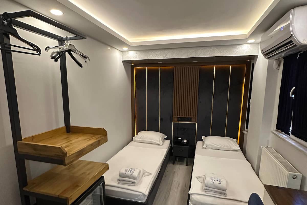 A compact hotel room with two single beds, a wooden shelf unit, a desk with a chair, and a built-in air conditioning unit.