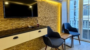 A modern living area with a flat-screen TV mounted on a textured wall, a wooden sideboard, two blue chairs, and a large window offering a view of the city. - Sirkeci Quietness Hotel (Istanbul)