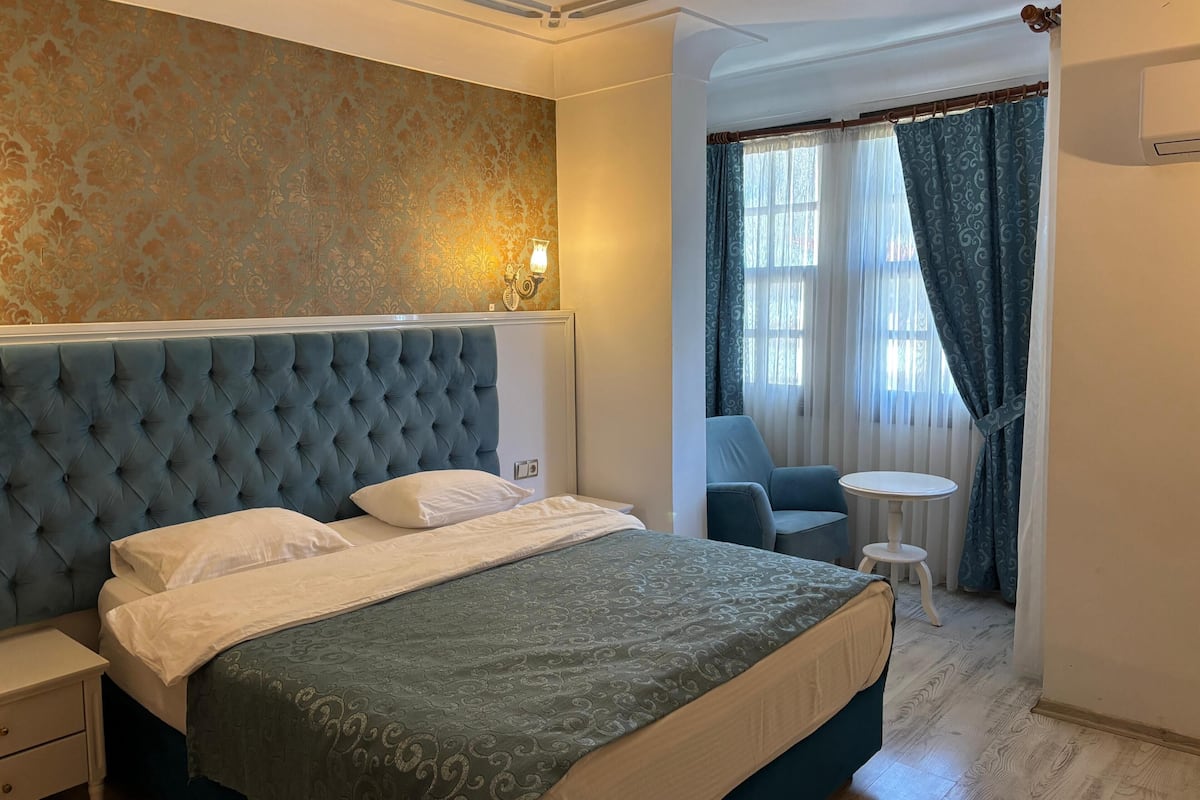 A bedroom with a tufted headboard, a bed with a patterned bedspread, a nightstand, a chair, a small round table, and a window with curtains.