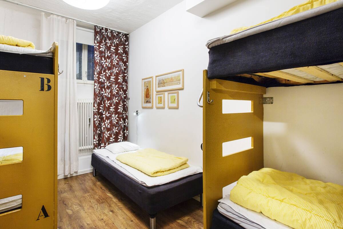 A dormitory room with bunk beds, a single bed, a radiator, and a window with curtains.