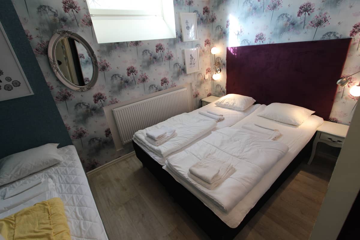 A double bed with white linens, a bedside table with a lamp, a mirror, and floral wallpaper.