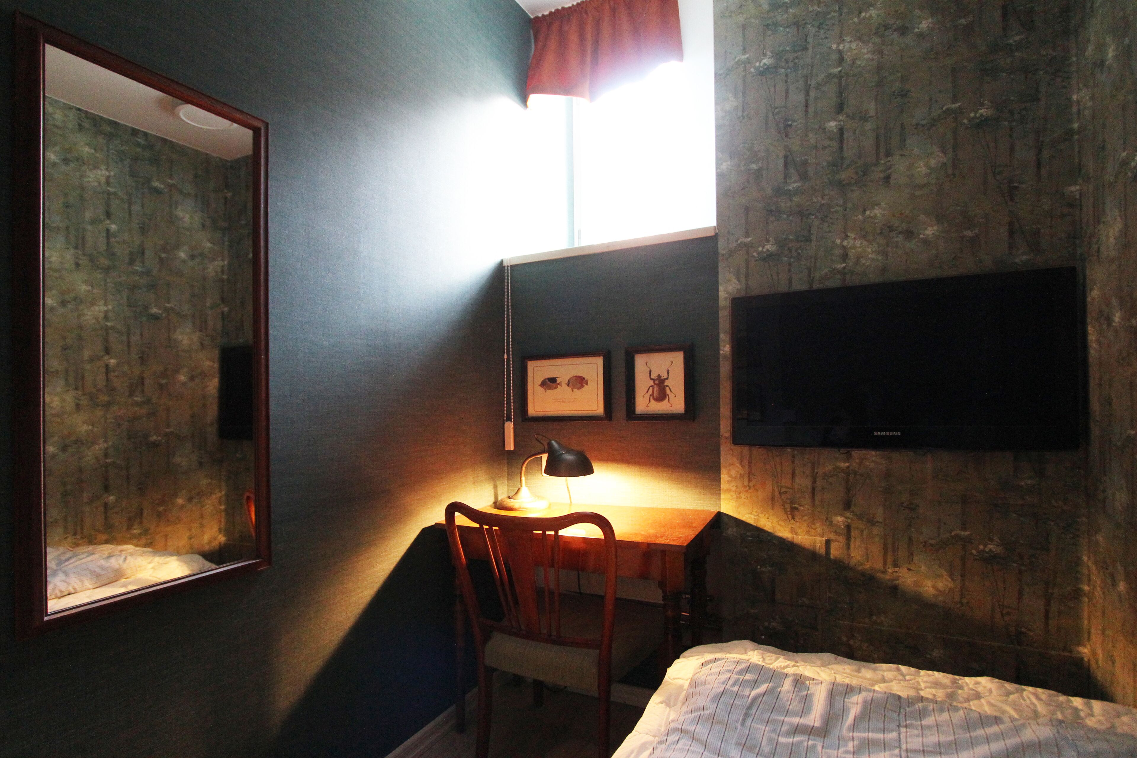 A bedroom with a bed, a desk with a chair, a mirror, and a wall-mounted TV.