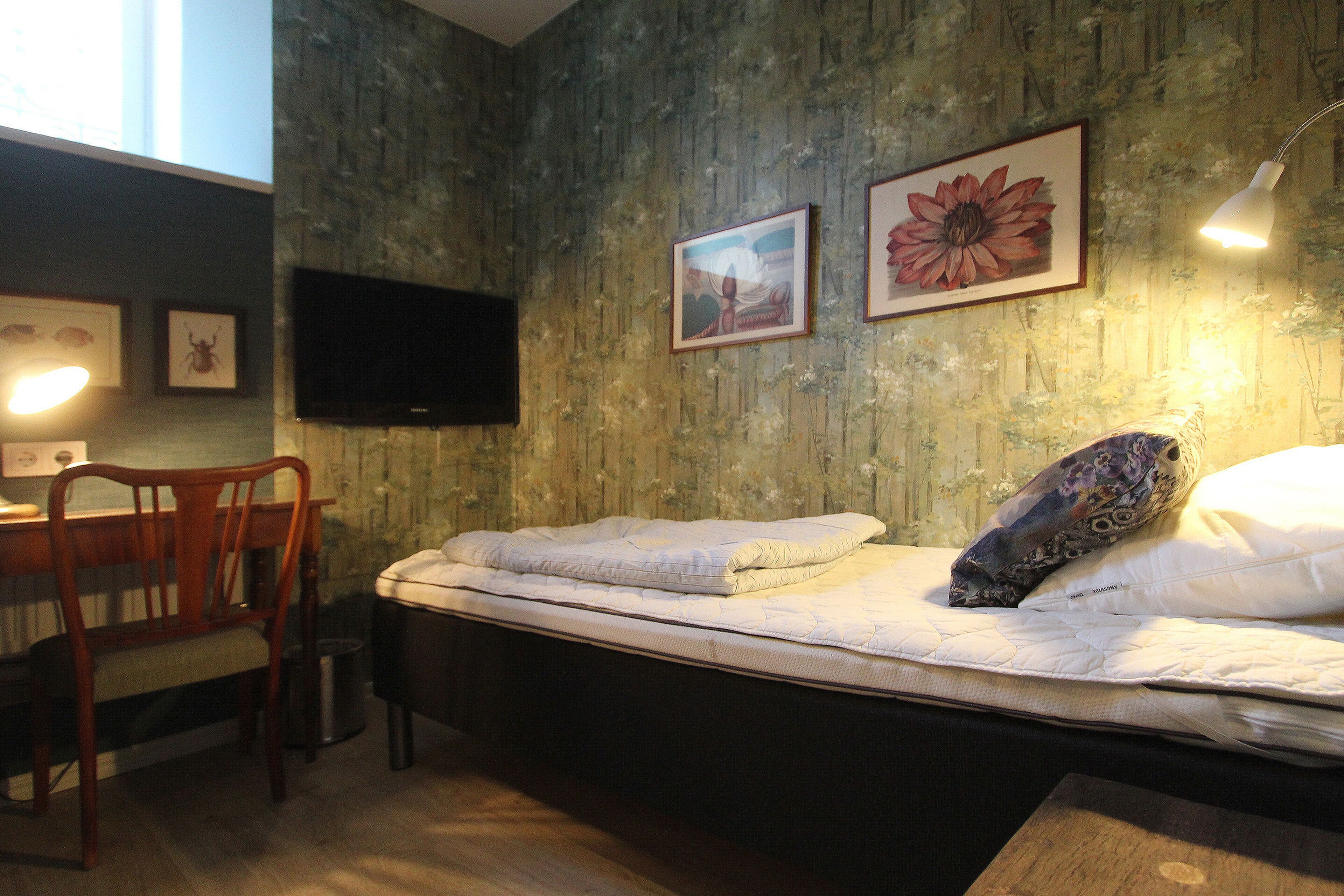 A bedroom with a bed, a desk, and a chair. There are framed pictures on the wall and a television above the desk.
