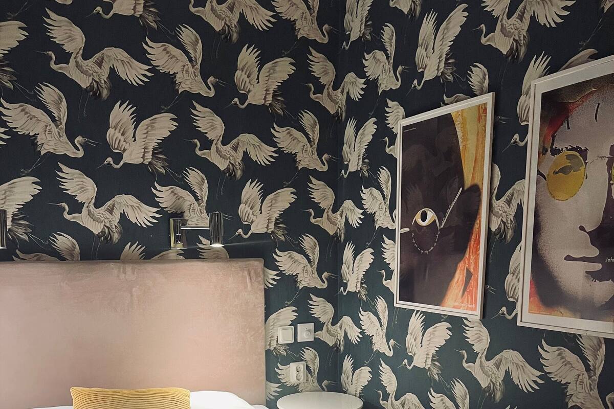 A neatly made bed with a yellow pillow, a bedside table with a lamp, and a wall with a bird pattern and a framed artwork.