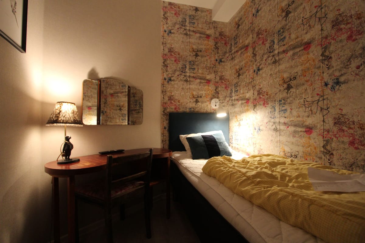 A small, dimly lit room with a bed, a desk, and a chair. The walls are covered with graffiti.