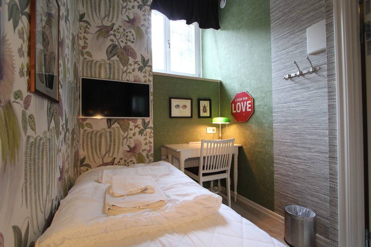 A room with a bed, a desk, a chair, a TV, a wall-mounted lamp, and a 'STOP FOR LOVE' sign.