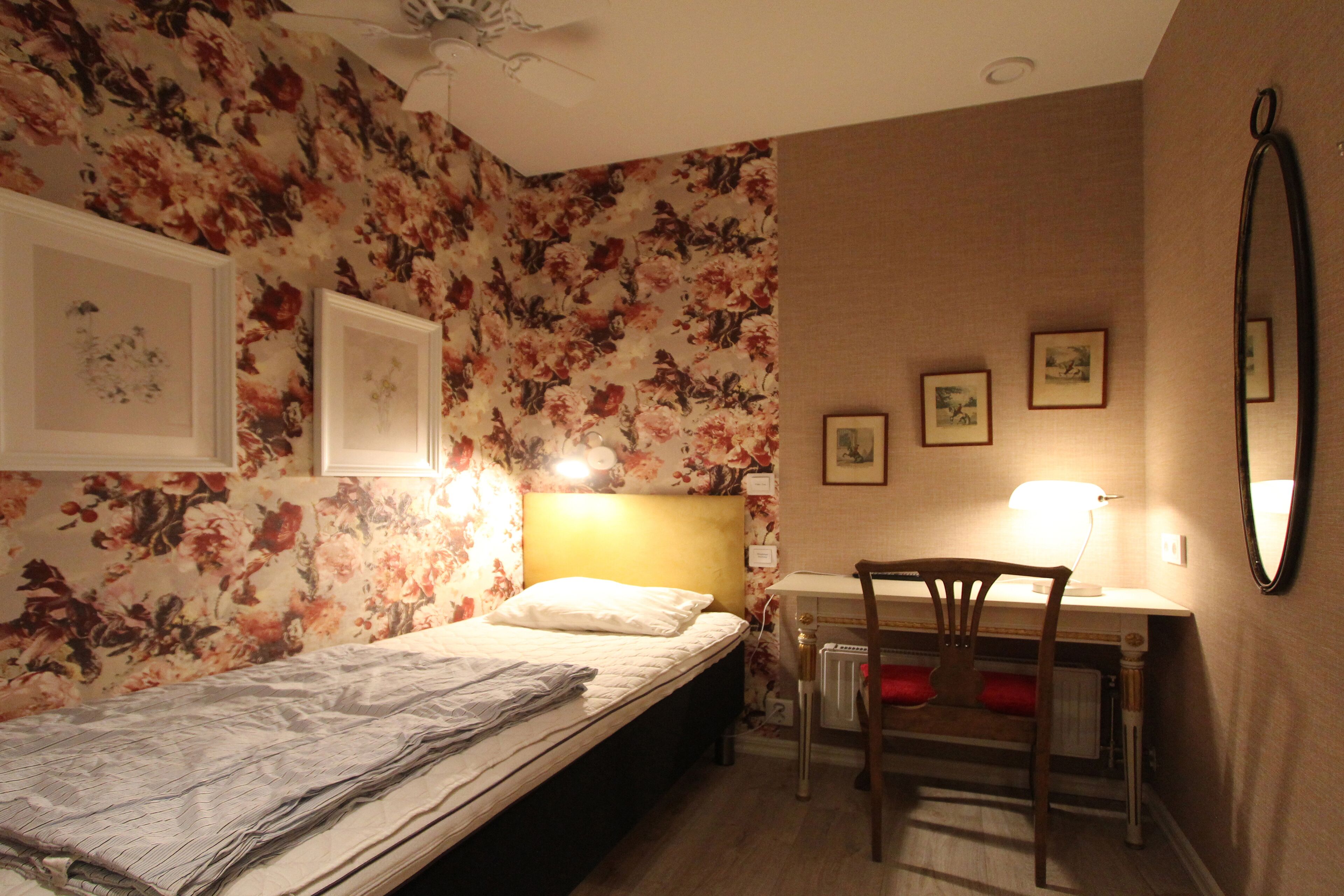 A bedroom with a bed, a desk, and a chair. There are floral wallpaper and framed pictures on the walls.