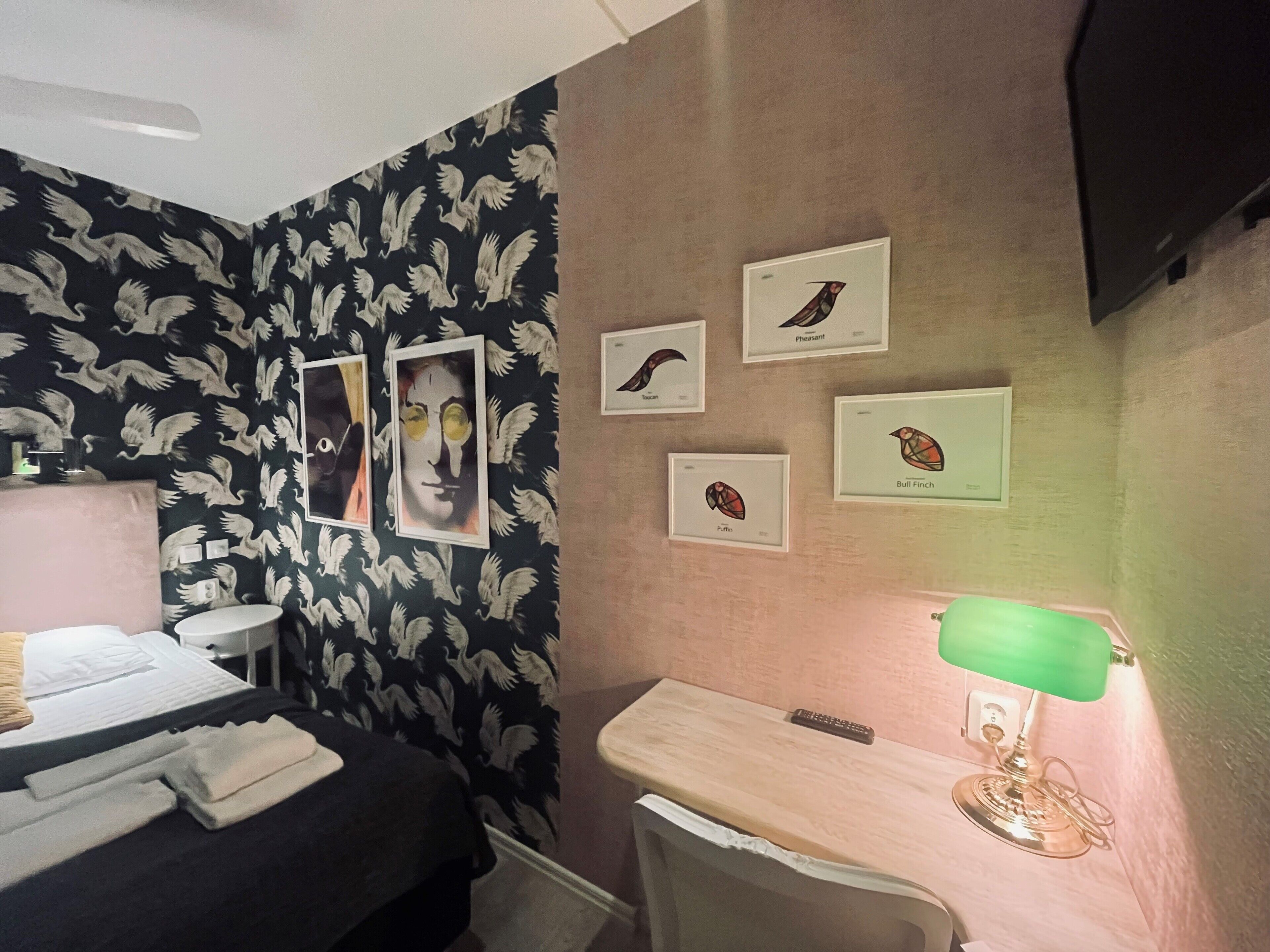 A hotel room with a bed, a desk, a chair, a green lamp, and wall art depicting birds.