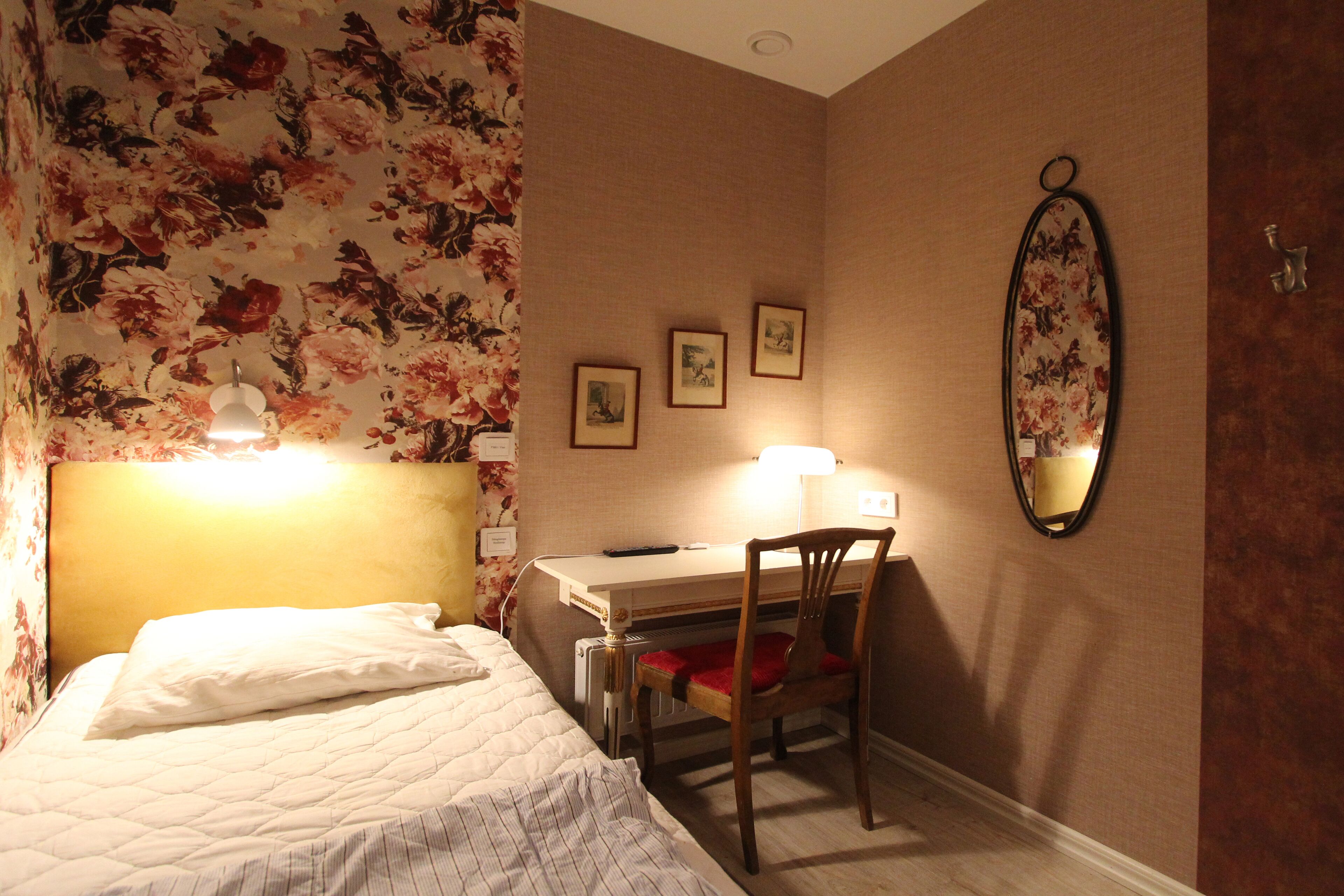 A hotel room with a bed, a desk with a chair, a wall-mounted mirror, and framed pictures.