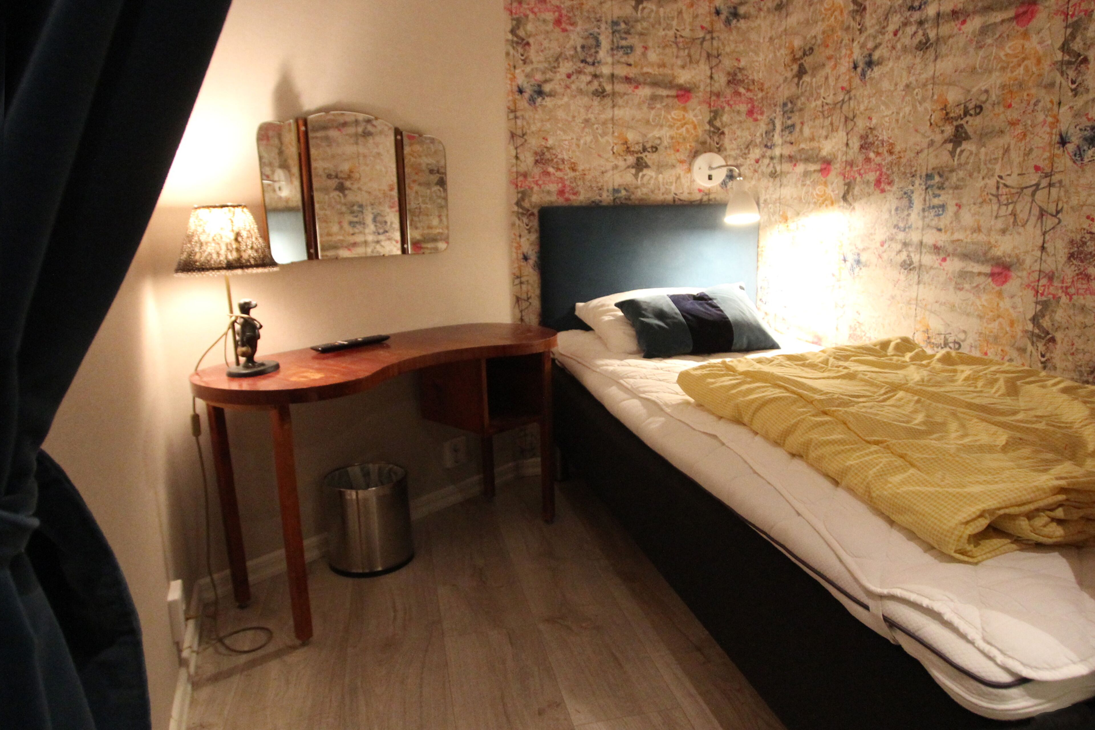 A bedroom with a bed, a desk, a lamp, and a wall with floral wallpaper.