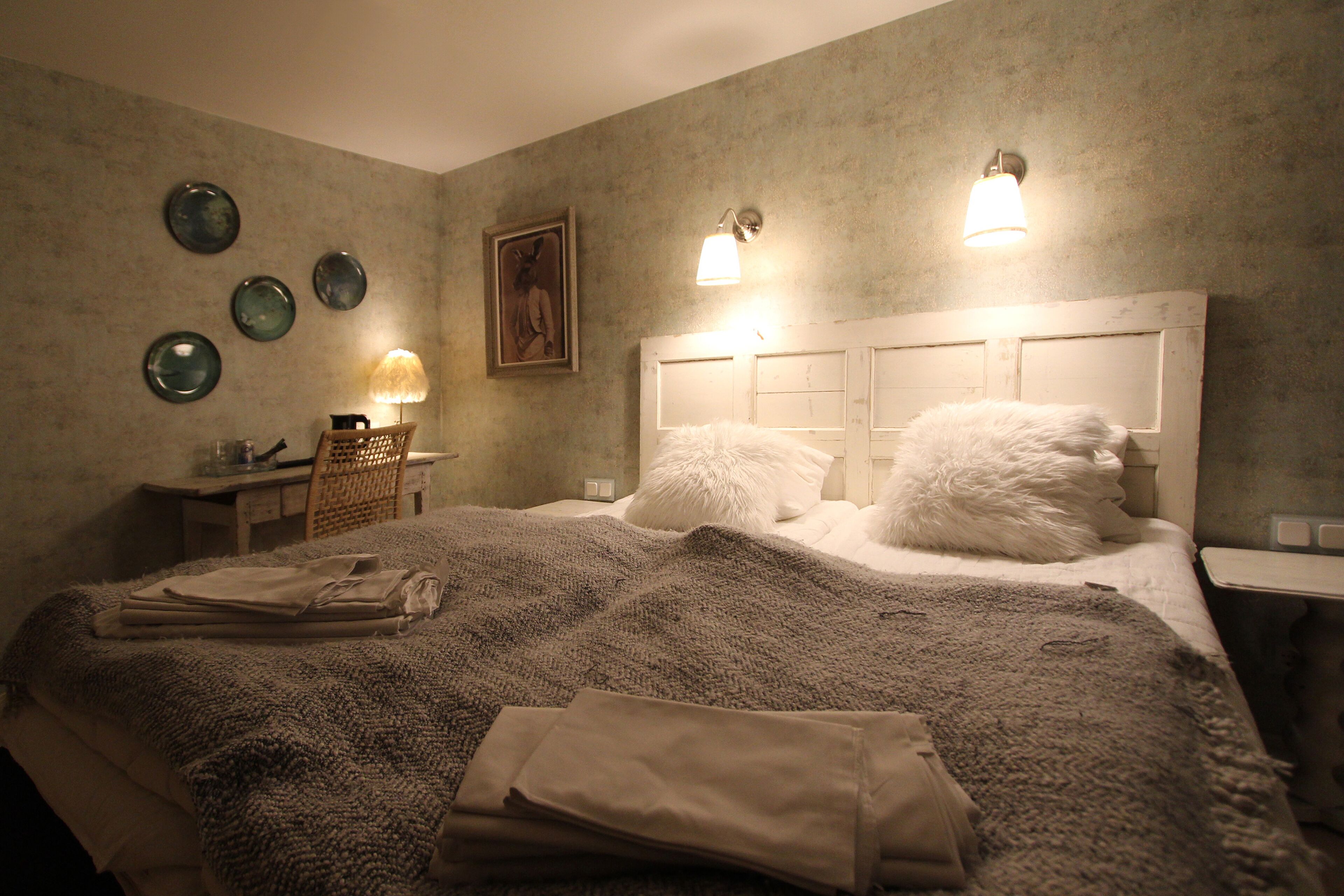 A bedroom with a large bed, a desk, and a chair. There are wall-mounted lights and a painting on the wall.