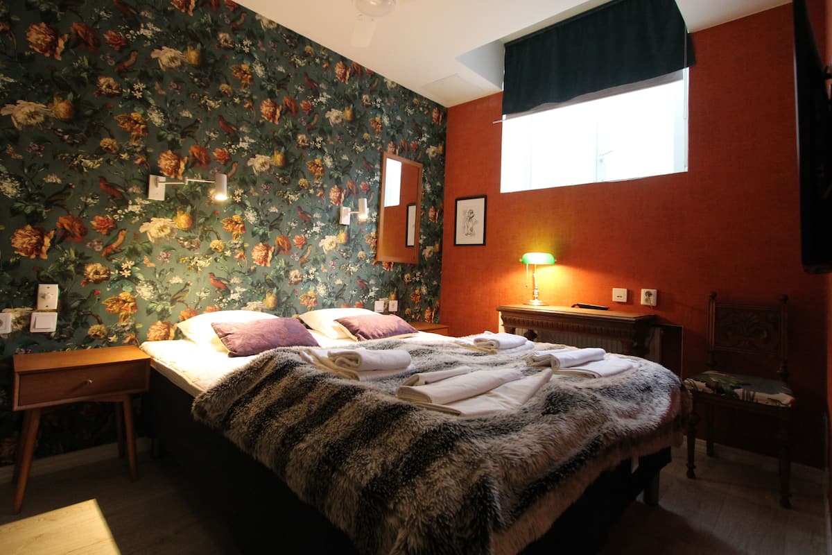 A bedroom with a floral wallpaper, a bed with a fur throw, a nightstand, a chair, and a window with a black valance.
