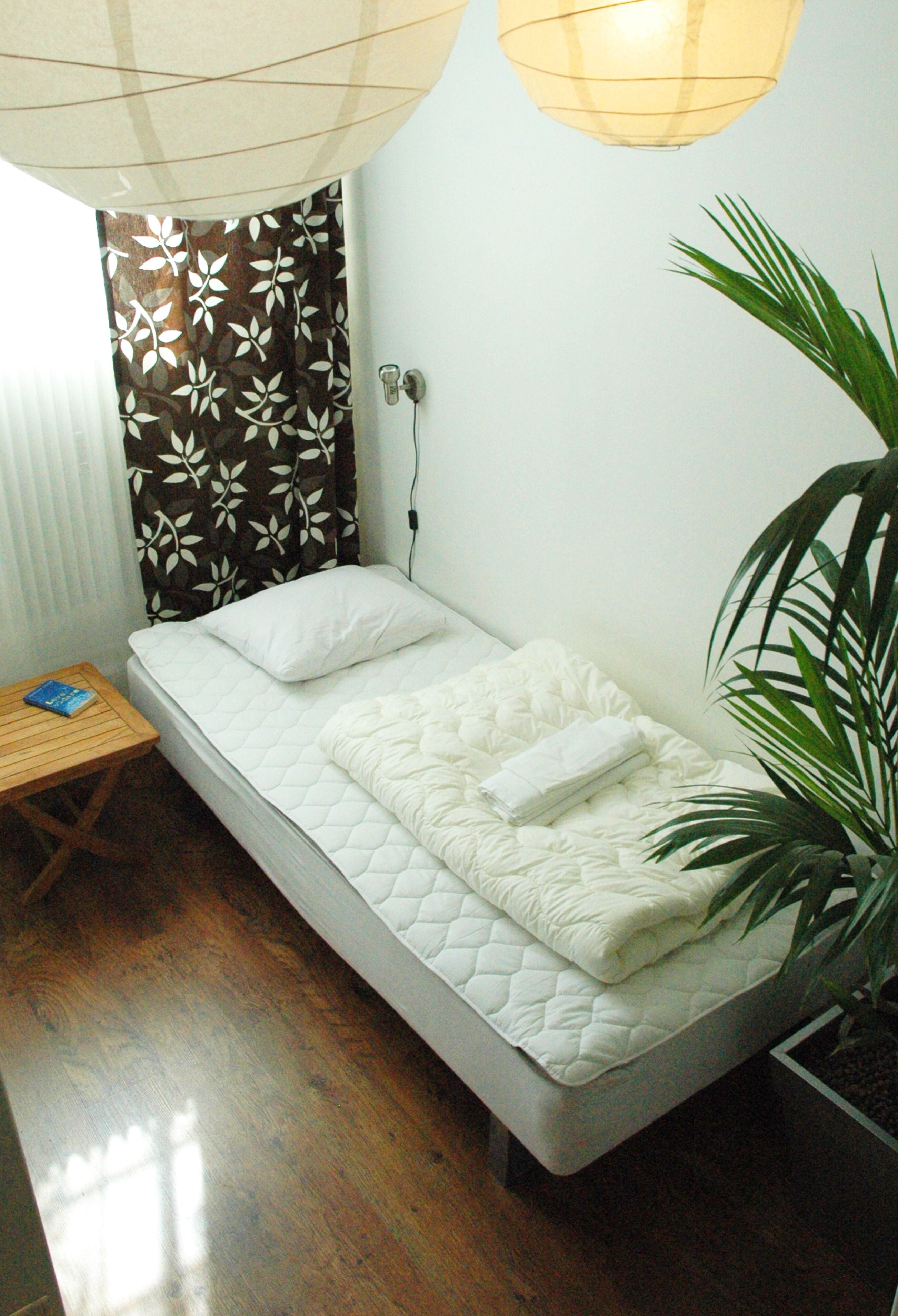 A single bed with a quilted mattress, a bedside table with a book, a potted plant, and a wall-mounted lamp.