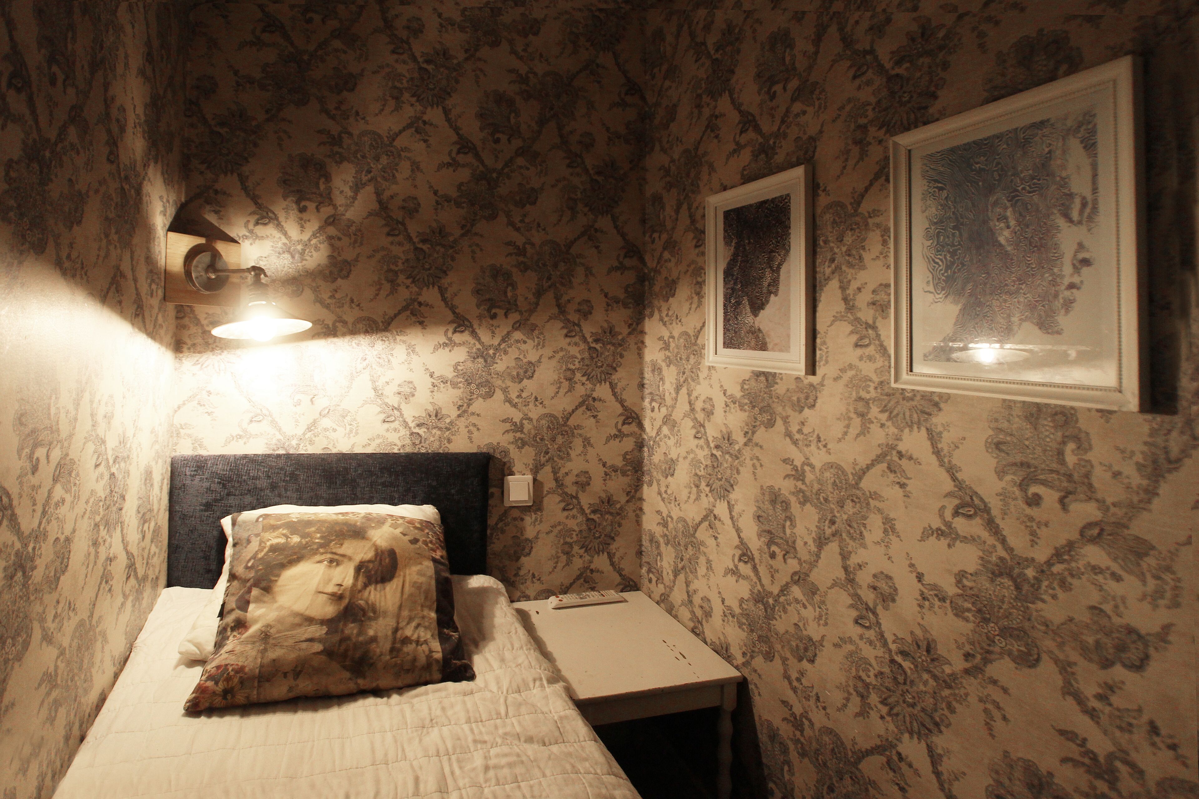 A bedroom with floral wallpaper, a bed with a pillow, a nightstand, and framed pictures on the wall.