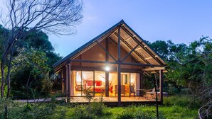 Une cabane moderne, dotée d’un toit triangulaire, avec une chambre à coucher aux murs de verre, une terrasse aménagée avec des sièges, et entourée d’une végétation luxuriante.