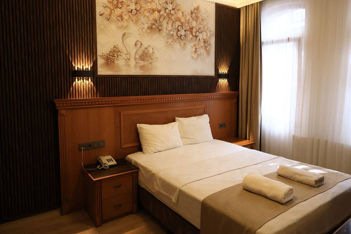 A hotel room with a large bed, wooden headboard, a nightstand with a phone, a wall-mounted light fixture, and a floral wall decoration.
