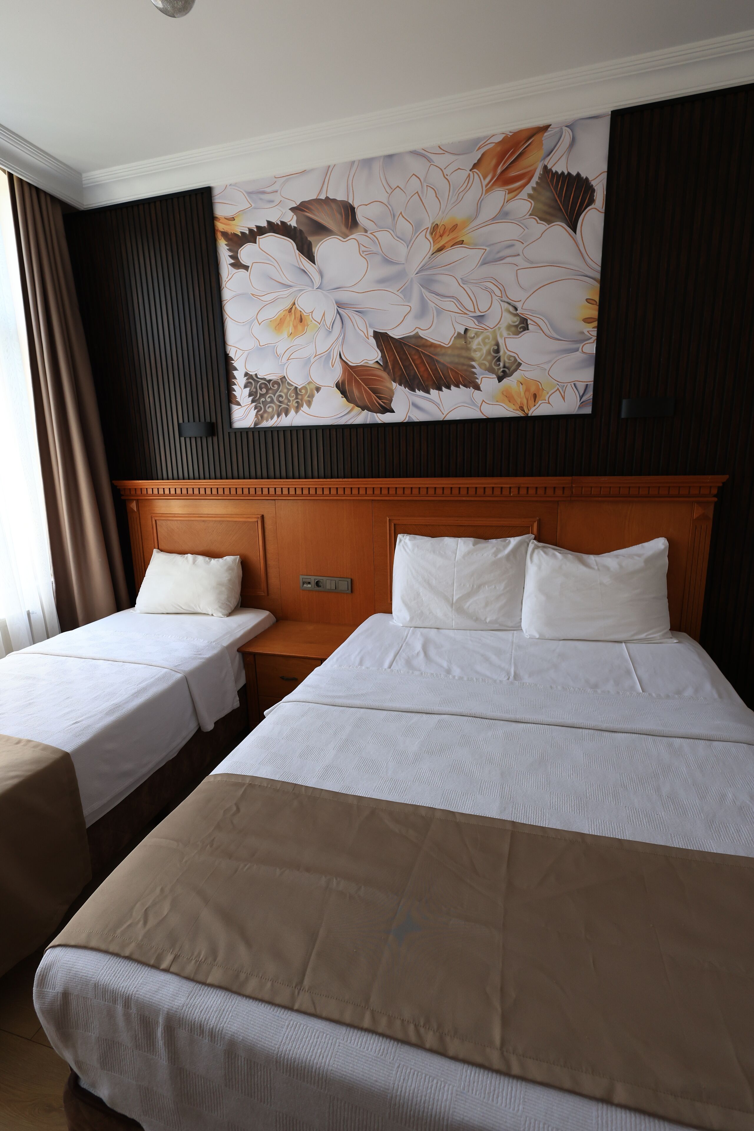 A hotel room with two single beds and one double bed, a wooden headboard, a large floral wall mural, and a window with curtains.