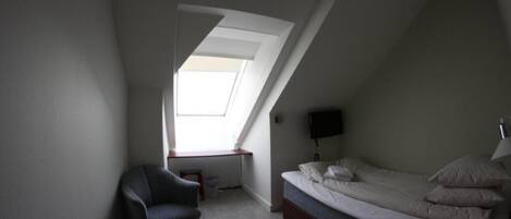 Una habitación pequeña con poca luz, techo inclinado, cama individual, una silla, escritorio y televisor fijado a la pared.