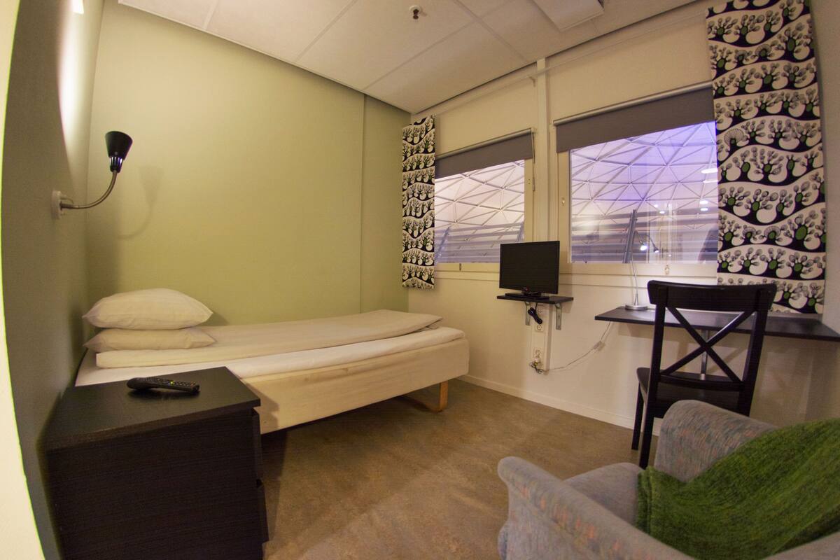 Une petite chambre avec un lit simple, un bureau et une chaise, une télévision et une fenêtre donnant sur un immeuble.
