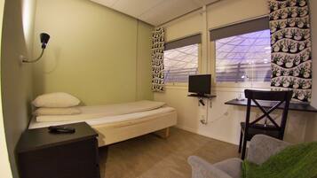 Une petite chambre avec un lit simple, un bureau et une chaise, une télévision et une fenêtre donnant sur un immeuble.