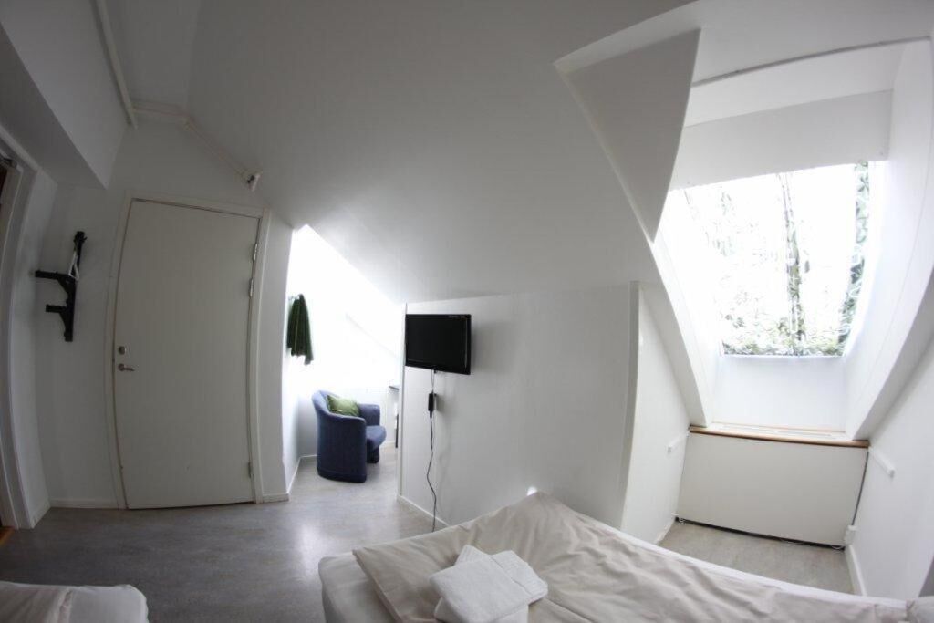 A modern, sparsely furnished room with a sloped ceiling, a door, a flat-screen TV mounted on the wall, and a window providing natural light.