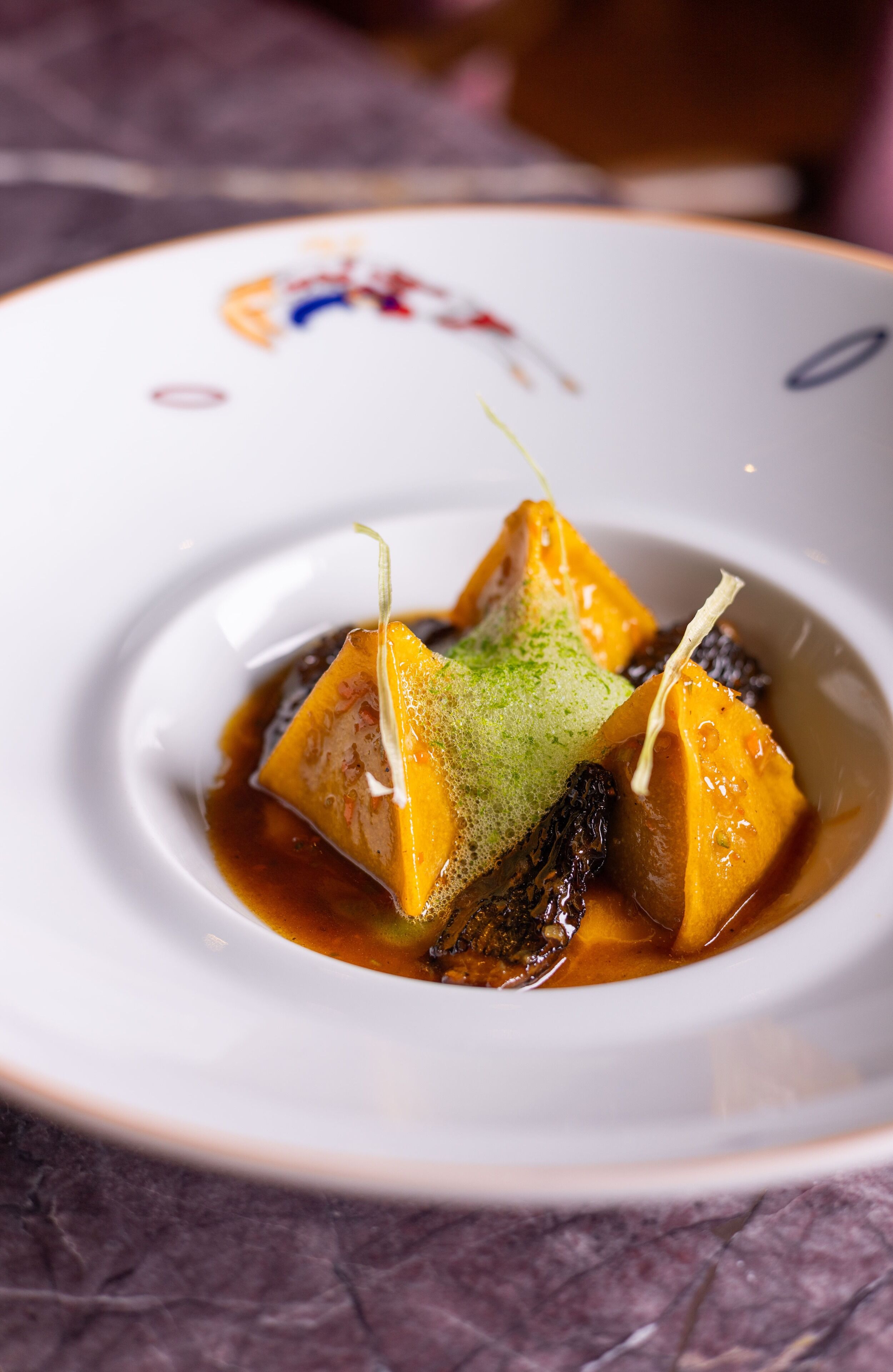 Une assiette blanche présente un dessert composé d’un cube d’aliments jaunes, peut-être du fromage, et d’une garniture verte, le tout servi dans une sauce foncée.