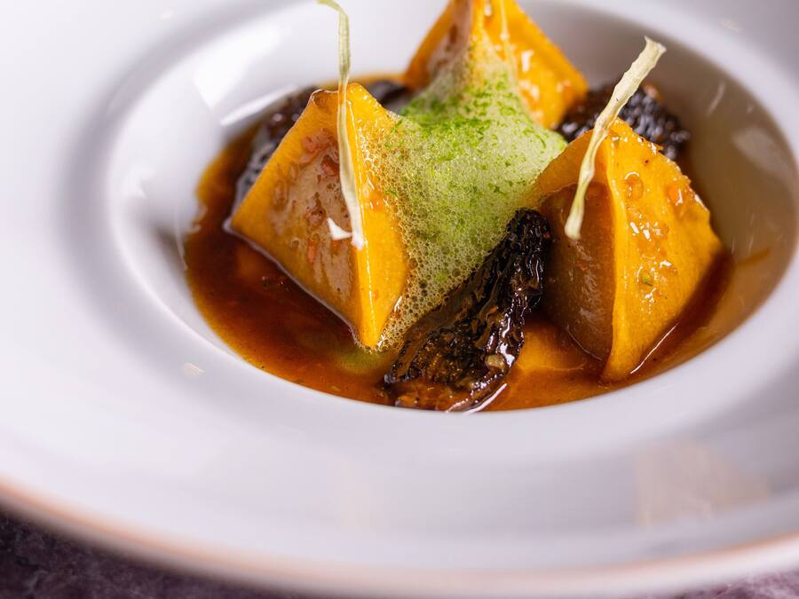A white plate with a dessert featuring a cube of yellow food, possibly cheese, and a green topping, served in a dark sauce.