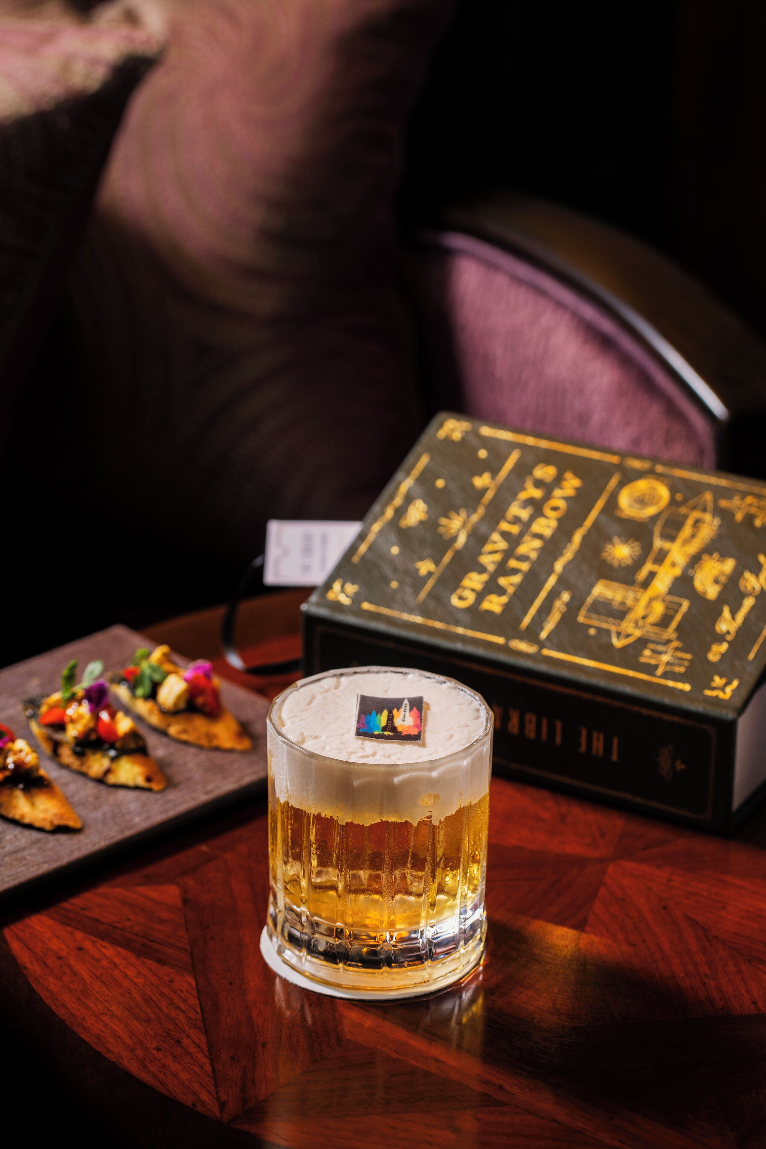 A glass of beer on a wooden table with a book titled "Gravity's Rainbow" in the background.