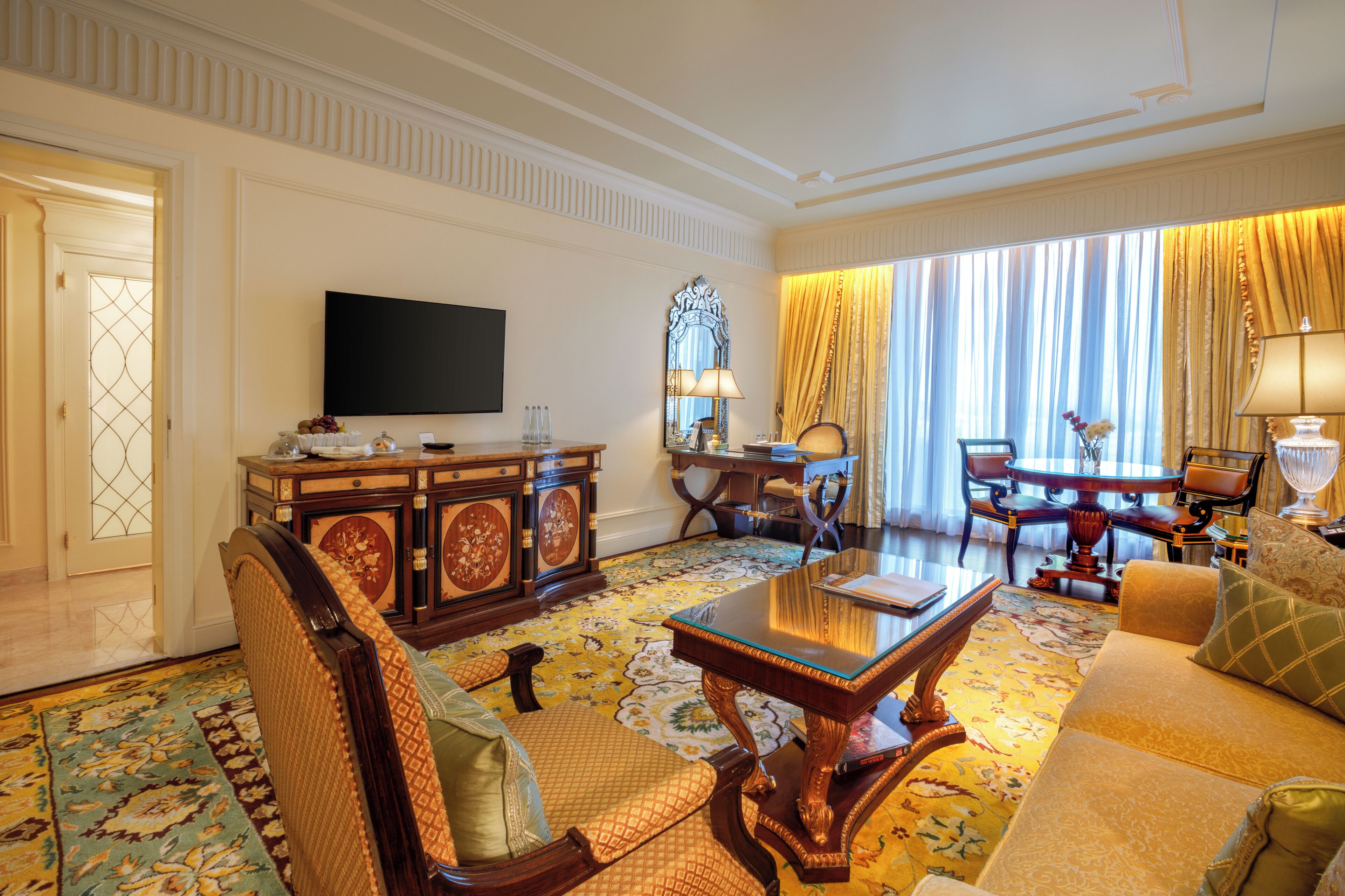 A spacious living room with a large sofa, a coffee table, and a TV. There is a dining table with chairs and a chandelier. The room has ornate wooden furniture and a patterned carpet.