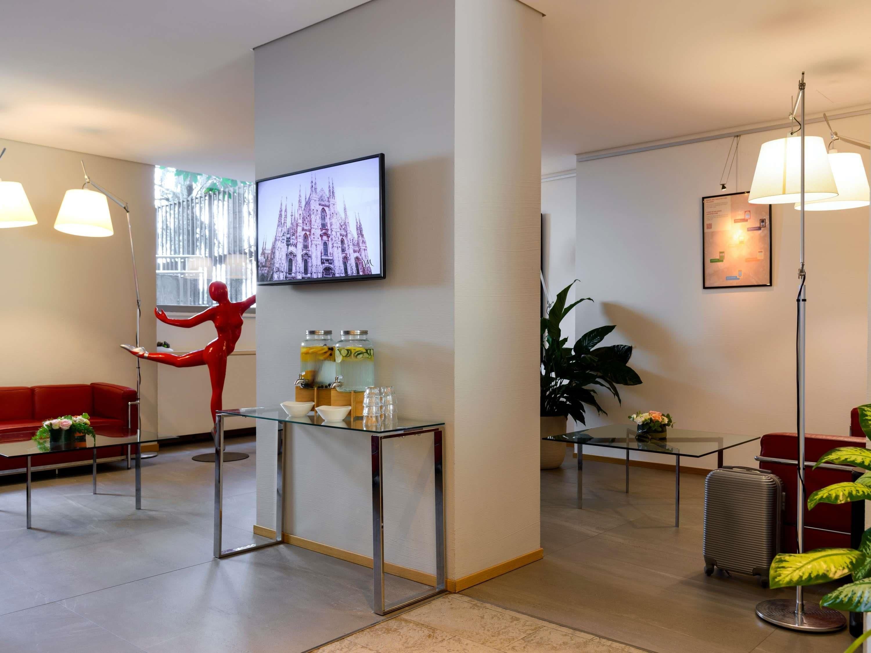 A hotel lobby with a red sofa, a glass table with water dispensers, a television displaying a cathedral, and a large plant.